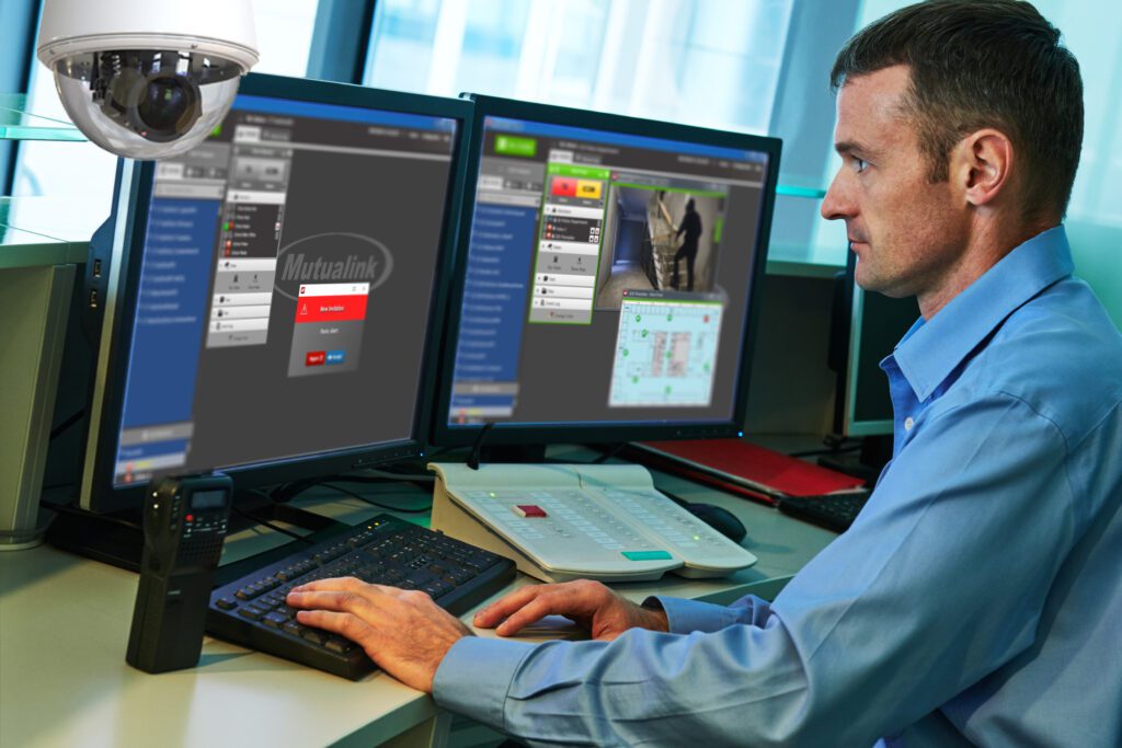 An emergency response officer is reviewing the details of an alert he just received. He is on the right hand side of the image looking to the left, where you can see his two monitors. On the monitors you see he is using Mutualink's Automated Emergency Response solution - the sophisticated and secure technology gives him immediate visual and communication access with the school who made the alert, as well as smart floor plans. He is serious, focused, and dedicated to conducting the most efficient emergency response.