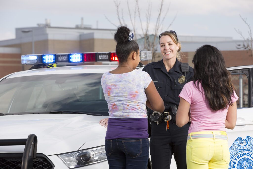 In the left hand side of the image you see the front of a police car. There is a school in the distance, visible in the top third of the image. The focus of the image, taking up the middle to right hand side, is a Police woman. She is facing the viewer, and appears relaxed, confident and happy. She is talking to two young girls, both of whom are facing the office, backs to us. You can tell everyone feels safe and comfortable. It appears that the office responded to an emergency at the school which was handled swiftly with a positive outcome. You can feel the joy and relief of all involved.