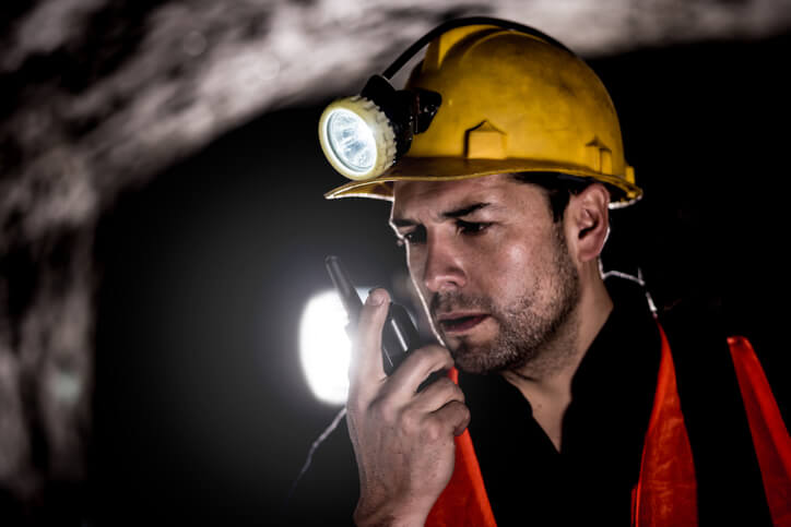 A man, underground in a mine, wears a hardhat with a light and has a slight grimace on his face holding a radio up to his mouth. He seems to be facing communication challenges in mines.